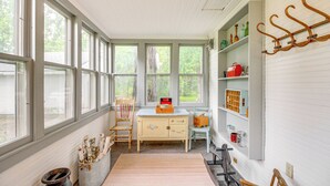 Een kamer met grote ramen, een houten stoel, een klein tafeltje en planken met diverse spullen.