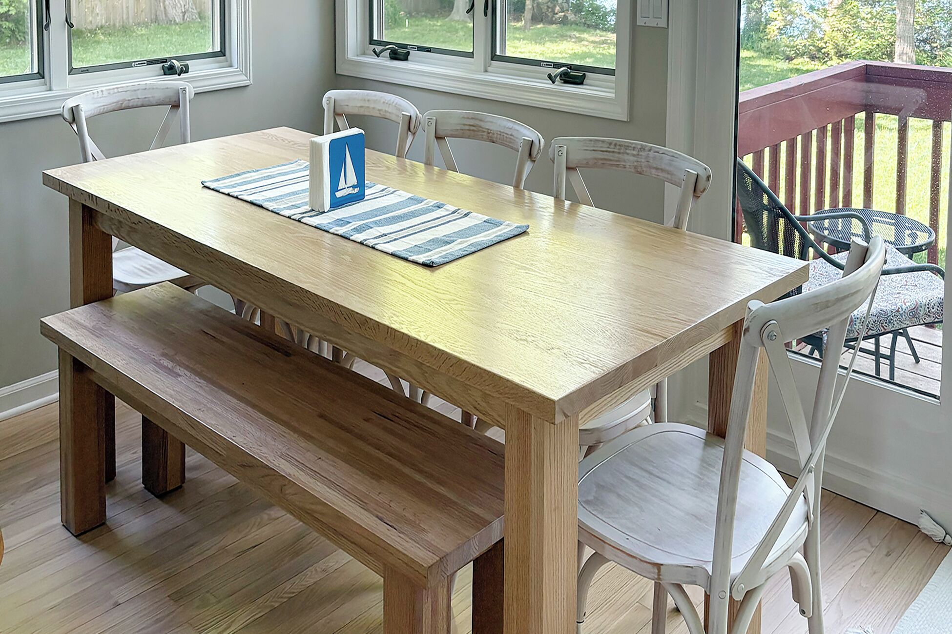 A wooden dining table with a bench, white chairs, and a view of a deck through a window.