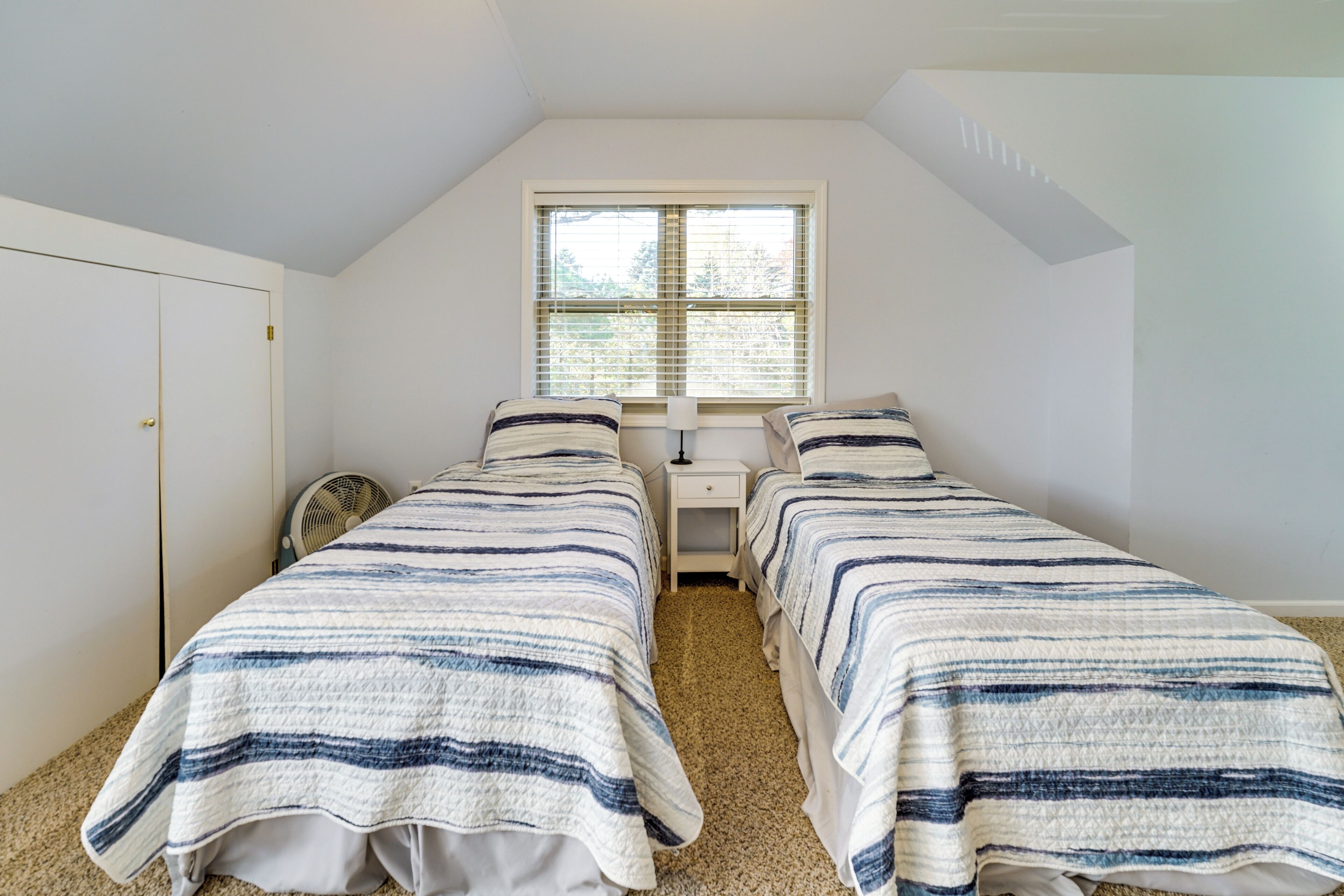 A bedroom with two single beds, a small bedside table with a lamp, a window with blinds, and a door.