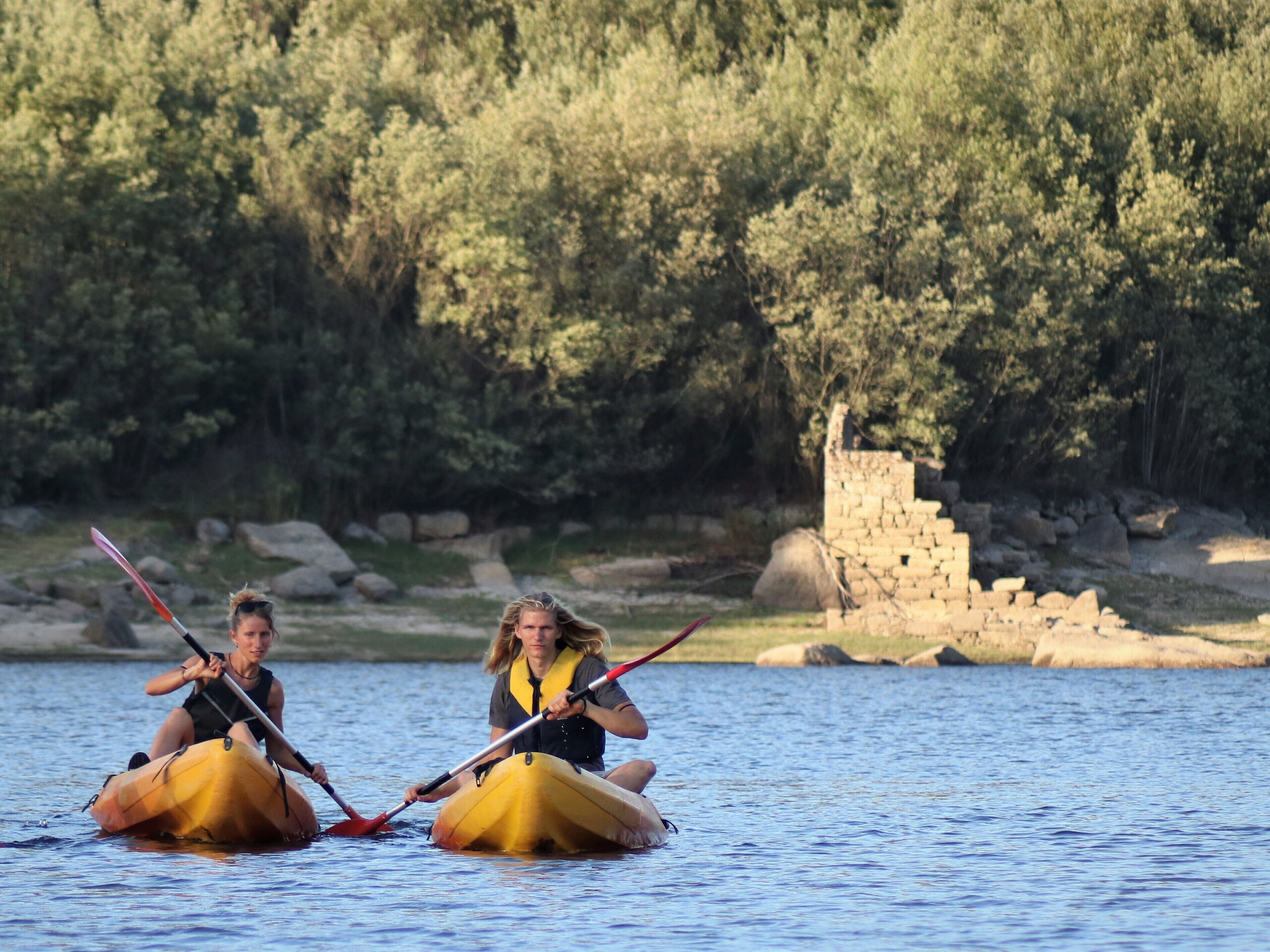 Deux personnes pratiquent le kayak sur une rivière paisible, avec une structure en pierre en arrière-plan.