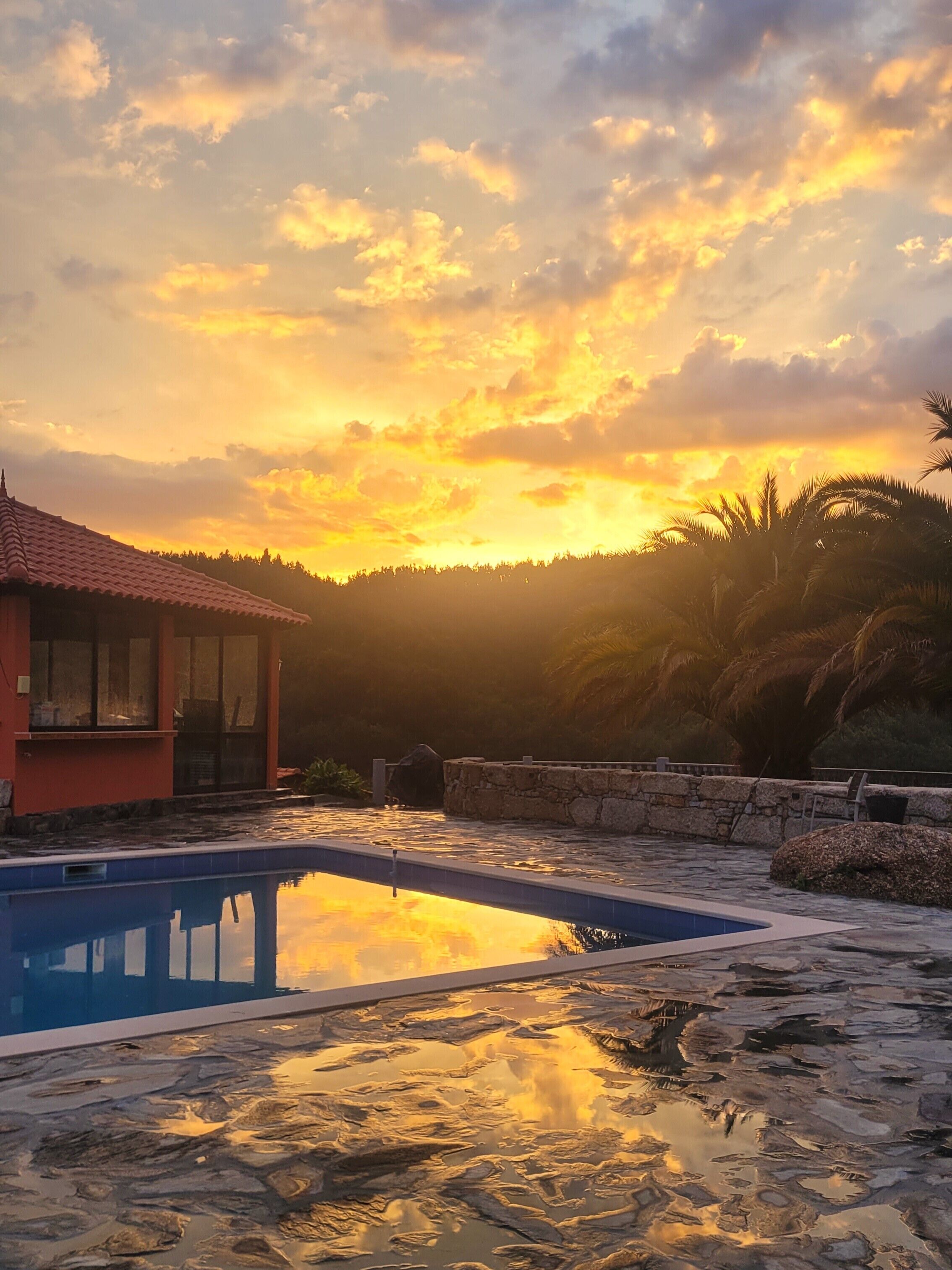 A swimming pool with a stone deck, a red-roofed building, and a sunset in the background.
