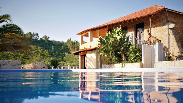 A poolside view of a stone building with a tiled roof, surrounded by lush greenery and a clear blue sky. - Encontro no Rio (Tábua)
