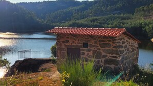 A stone house with a tiled roof near a lake, surrounded by greenery and hills. - Encontro no Rio (Tábua)