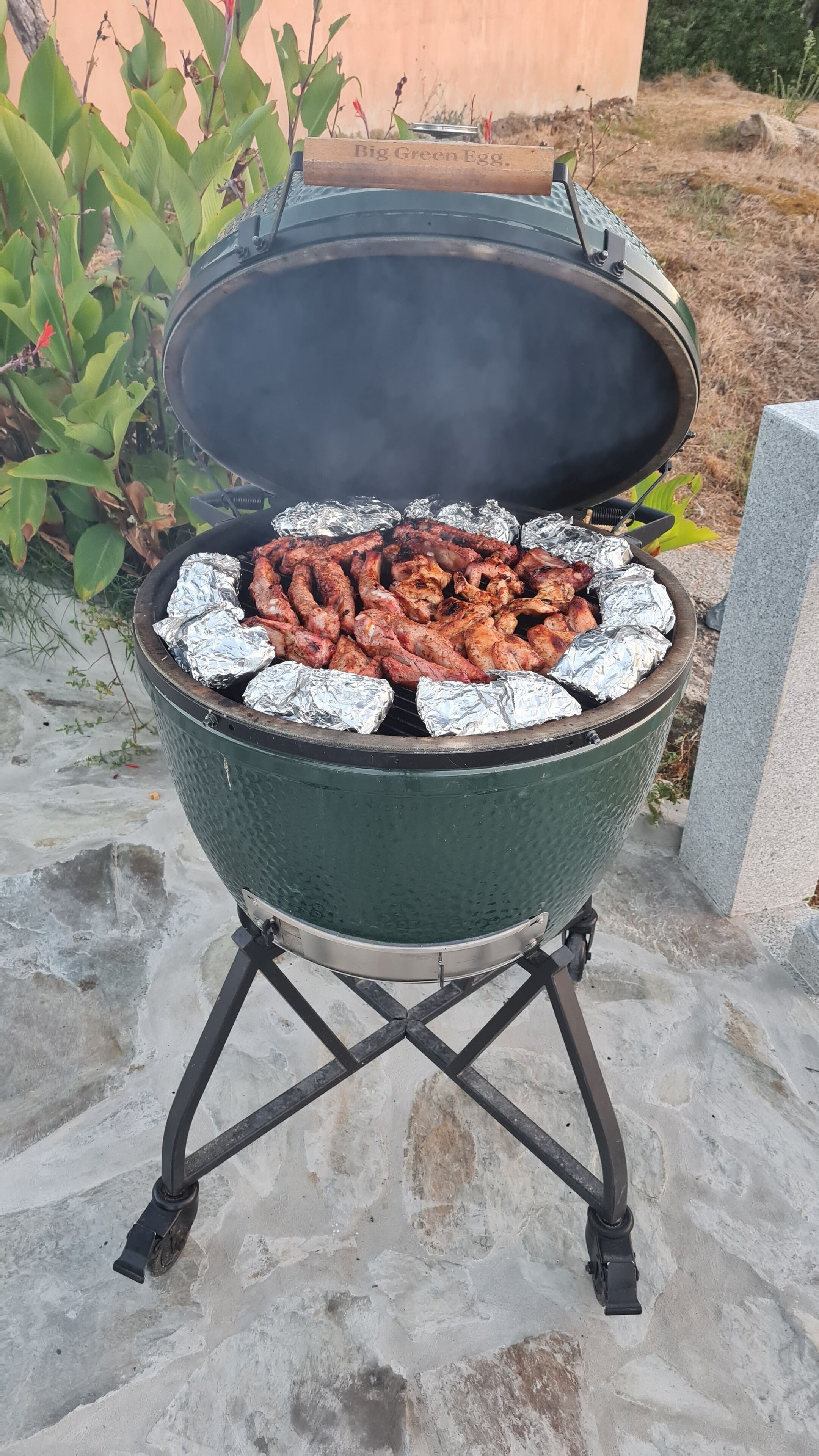 A portable grill with foil-wrapped meat, set outdoors on a stone surface.