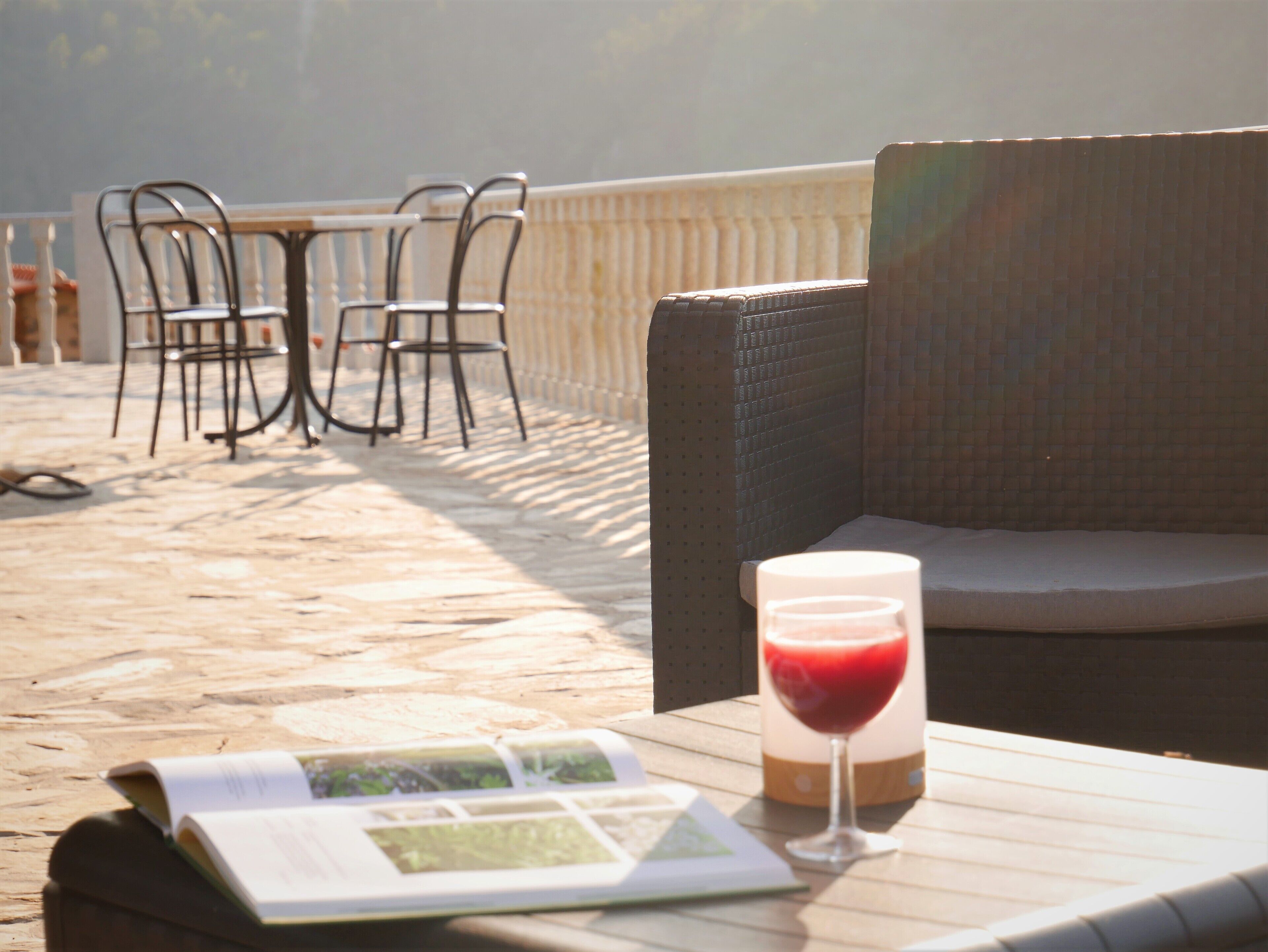 Ein Tisch mit einem Buch und einem Glas roter Flüssigkeit, Gartenmöbel aus Rattan und ein Fliesenboden.