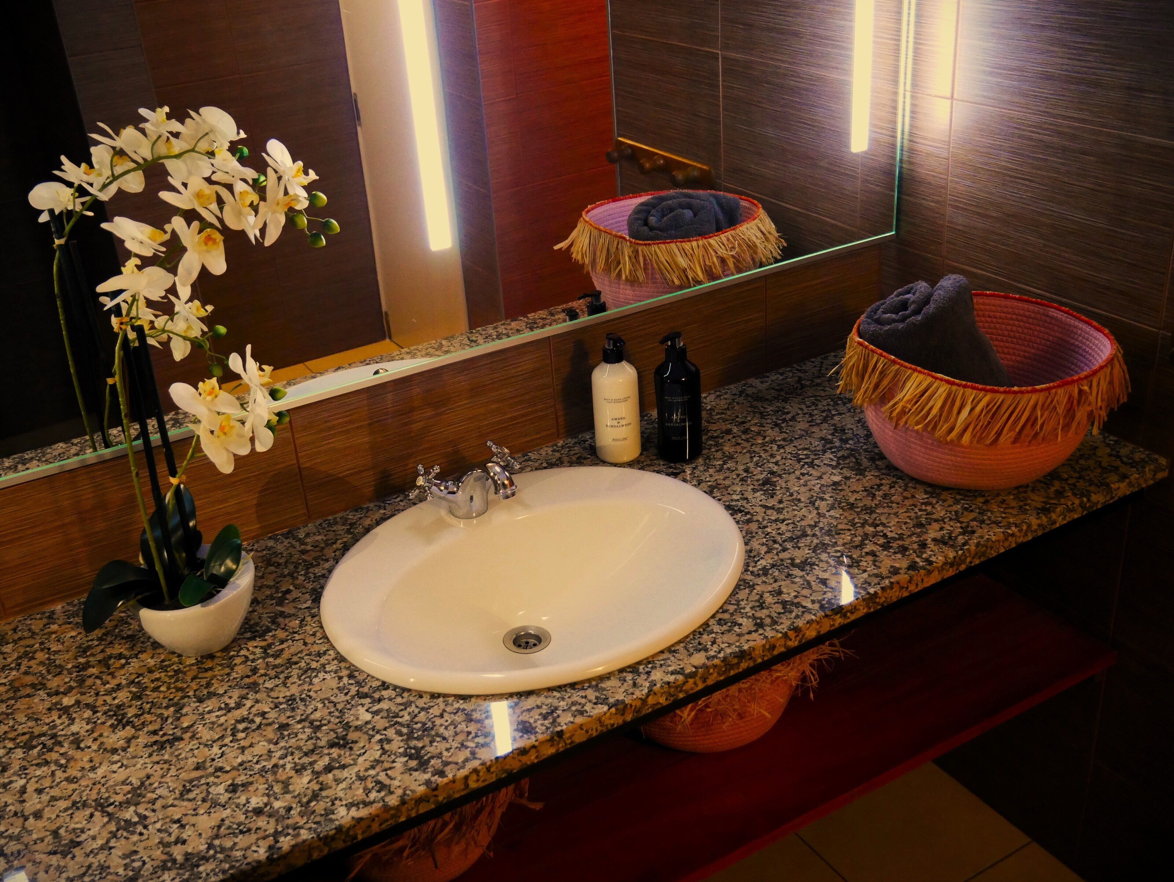 A bathroom with a granite countertop, white sink, and a mirror reflecting the room. There are two bottles on the counter and a woven basket with a towel.