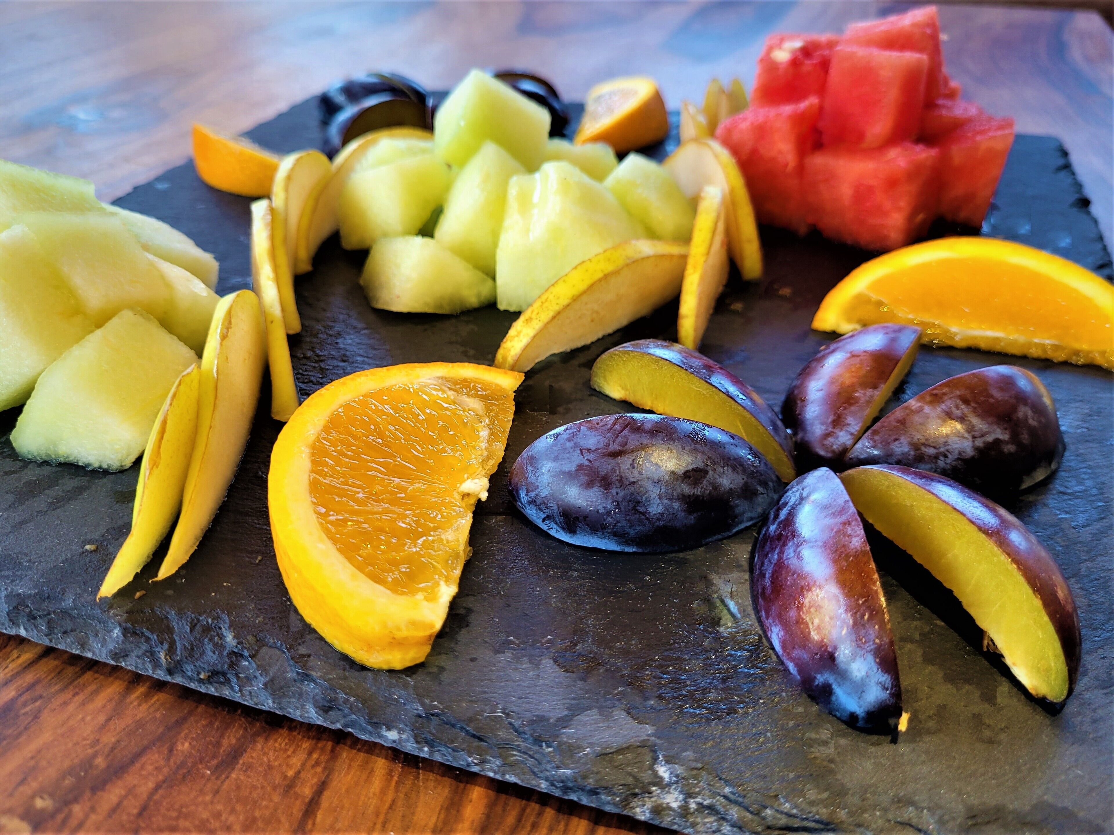 A variety of fruits including watermelon, cantaloupe, orange, and plums arranged on a slate board.