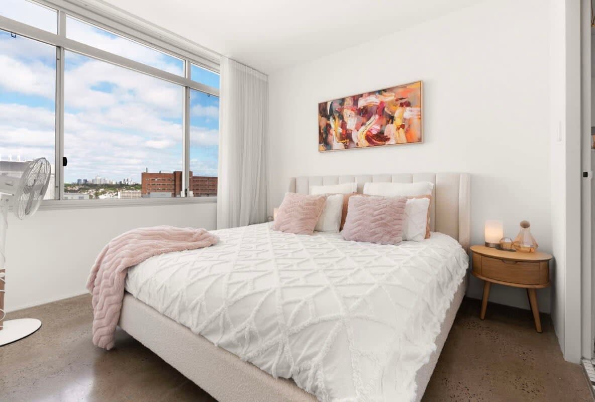 A bedroom with a large bed, a bedside table with a lamp, a window with a city view, and a painting on the wall.