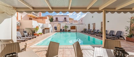A covered outdoor swimming pool with lounge chairs and tables, surrounded by buildings.