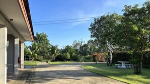 A quiet street with a building on the left, a grassy area with trees, and a clear sky. - Baanmae Resort Thachana Suratthani (Tha Chana)