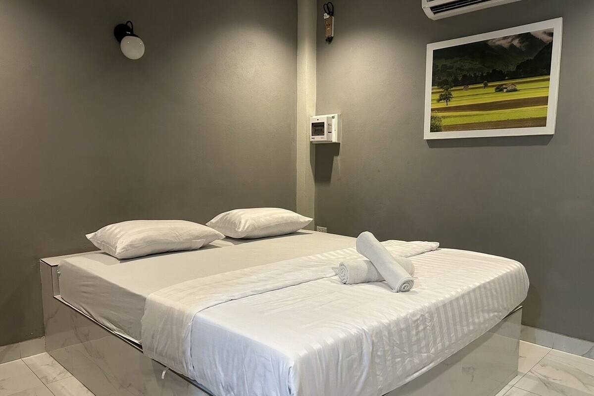 A neatly made bed with white linens and pillows, a small bedside table, and a wall-mounted air conditioner.