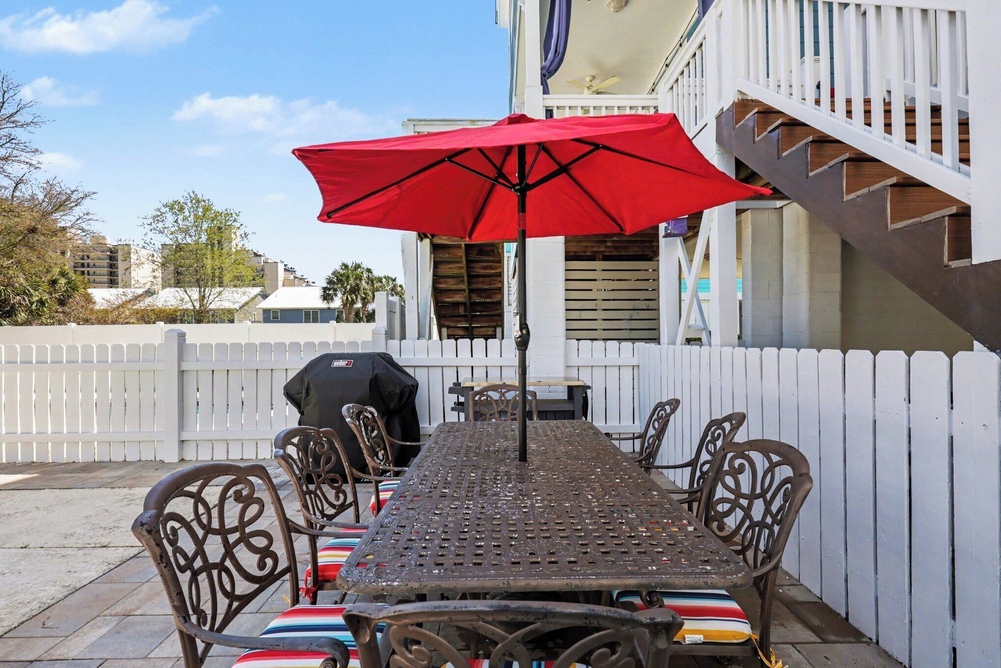 Une terrasse avec une table et des chaises en métal, un parasol rouge et une clôture blanche en bois.