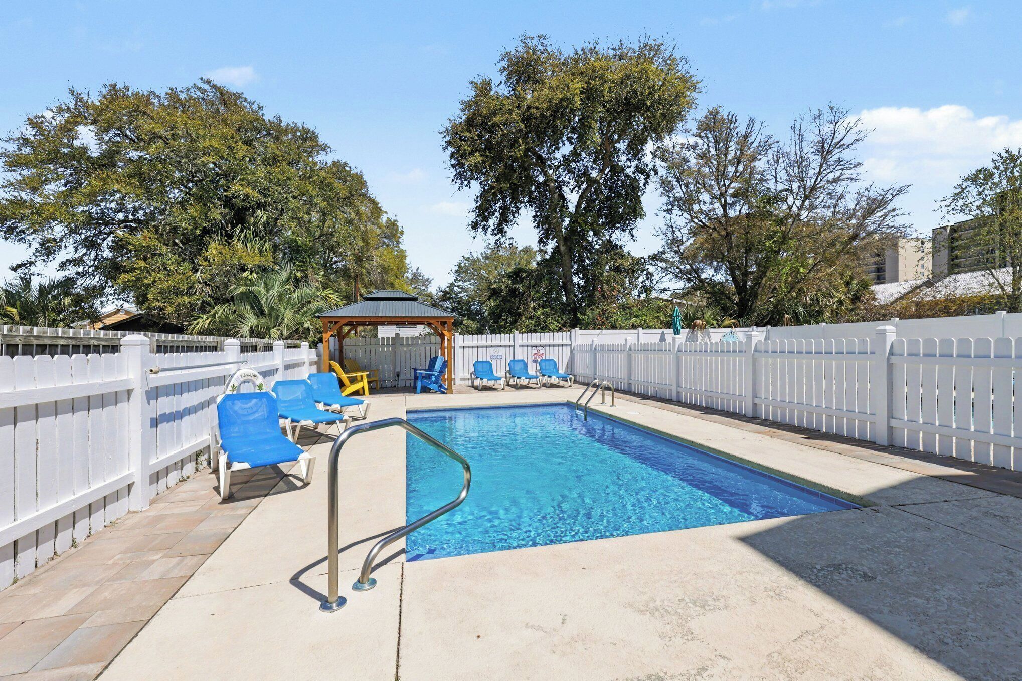 Une piscine rectangulaire avec un escalier en métal, entourée d’une clôture blanche et agrémentée de chaises bleues.