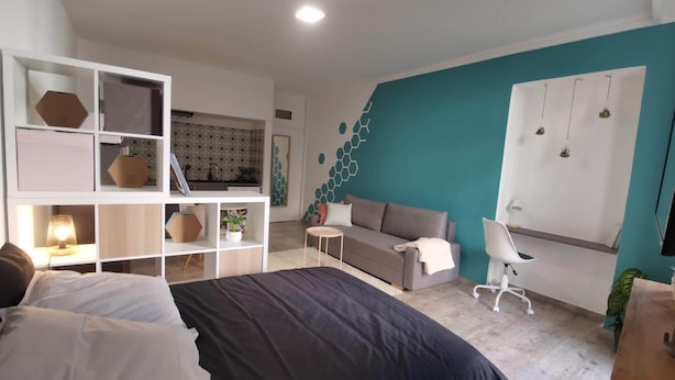 Une chambre moderne avec un lit, un canapé gris, un bureau et une chaise. On y trouve une étagère blanche agrémentée d’objets décoratifs, ainsi qu’un mur orné d’un motif géométrique.
