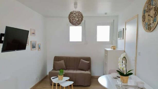 A modern living room with a brown sofa, a small white table with two chairs, a TV mounted on the wall, and a decorative clock. - T1 - Studio Confortable Wissant (Wissant)