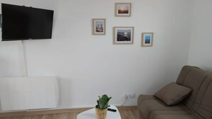 A living room with a flat-screen TV mounted on the wall, a brown sofa, a small white table with a plant, and framed pictures on the wall. - T1 - Studio Confortable Wissant (Wissant)