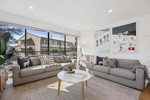 Ein modernes Wohnzimmer mit zwei grauen Sofas, einem weißen Couchtisch und großen Fenstern mit Blick auf ein Gebäude und Bäume.