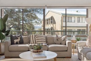 Una sala de estar moderna con sofá, mesa de centro y ventanas amplias que ofrecen vistas a edificios y árboles.