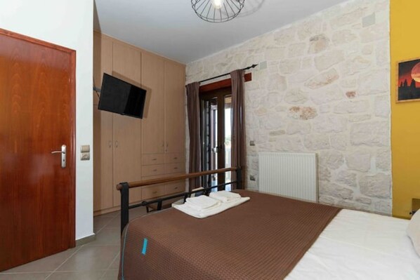 A bedroom with a bed, a television mounted on the wall, a wooden wardrobe, and a stone wall.