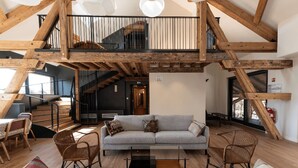 Een ruime loft met zichtbare houten balken, een trap, een grijze bank, rieten stoelen en een glazen salontafel.