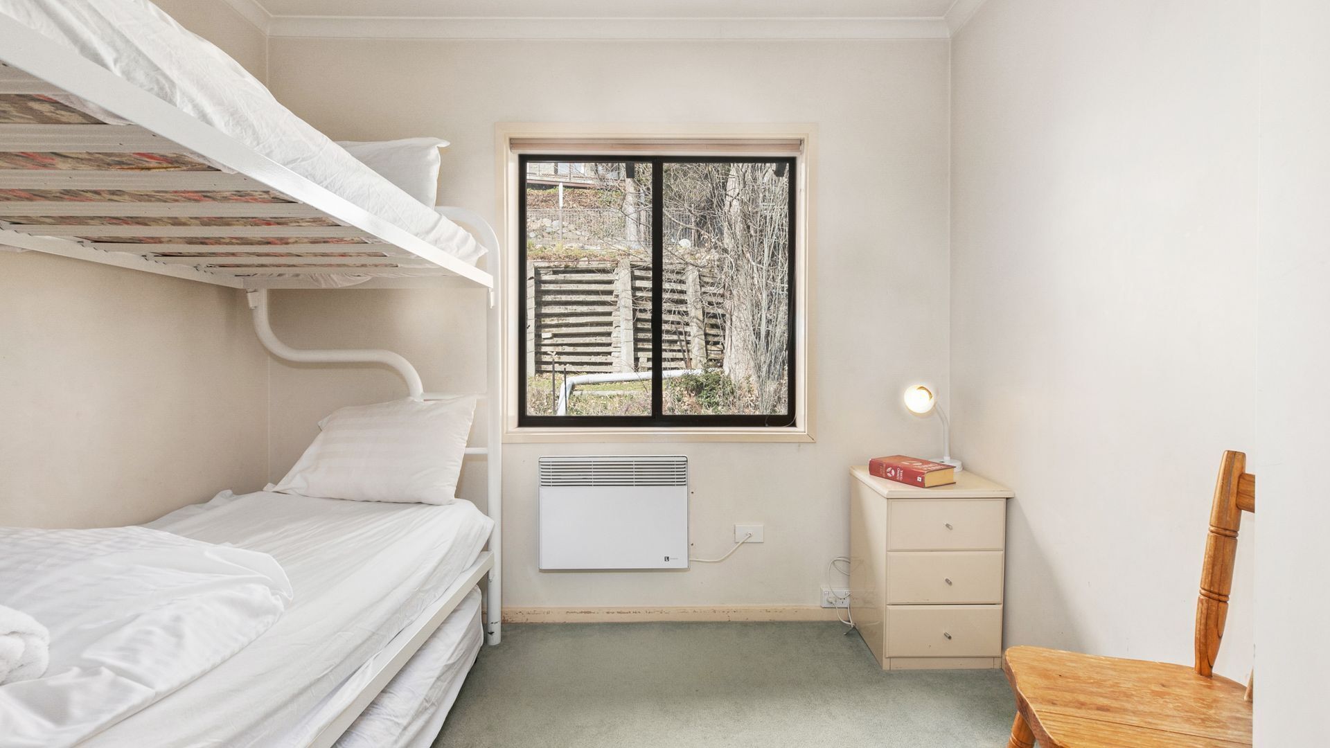 Una habitación pequeña y bien iluminada con literas, una ventana con vistas a edificios, una mesita de noche con lámpara y una silla.