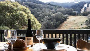 Una mesa con una botella de vino, copas y un bol de comida en un balcón con vistas a un paisaje montañoso.