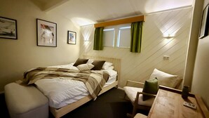 Een slaapkamer met een bed, een stoel, een klein tafeltje en een raam met gordijnen.