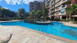 Piscina com borda de pedra, rodeada por espreguiçadeiras e palmeiras, com um edifício de vários andares ao fundo.