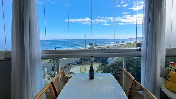 Mesa com uma garrafa de vinho Valdo e duas taças, com vista para uma praia e paisagem urbana através de grandes janelas.