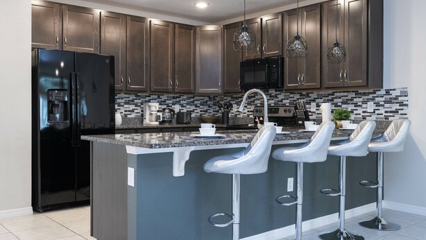 A modern kitchen with dark cabinets, stainless steel appliances, a black refrigerator, a microwave, and a bar area with white bar stools.