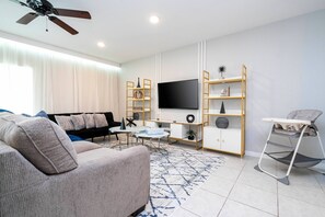 Experience a bright and airy living room with a plush gray sofa and stylish area rug, perfect for cozy gatherings. Enjoy the modern entertainment setup and natural light from large windows to enhance your stay.