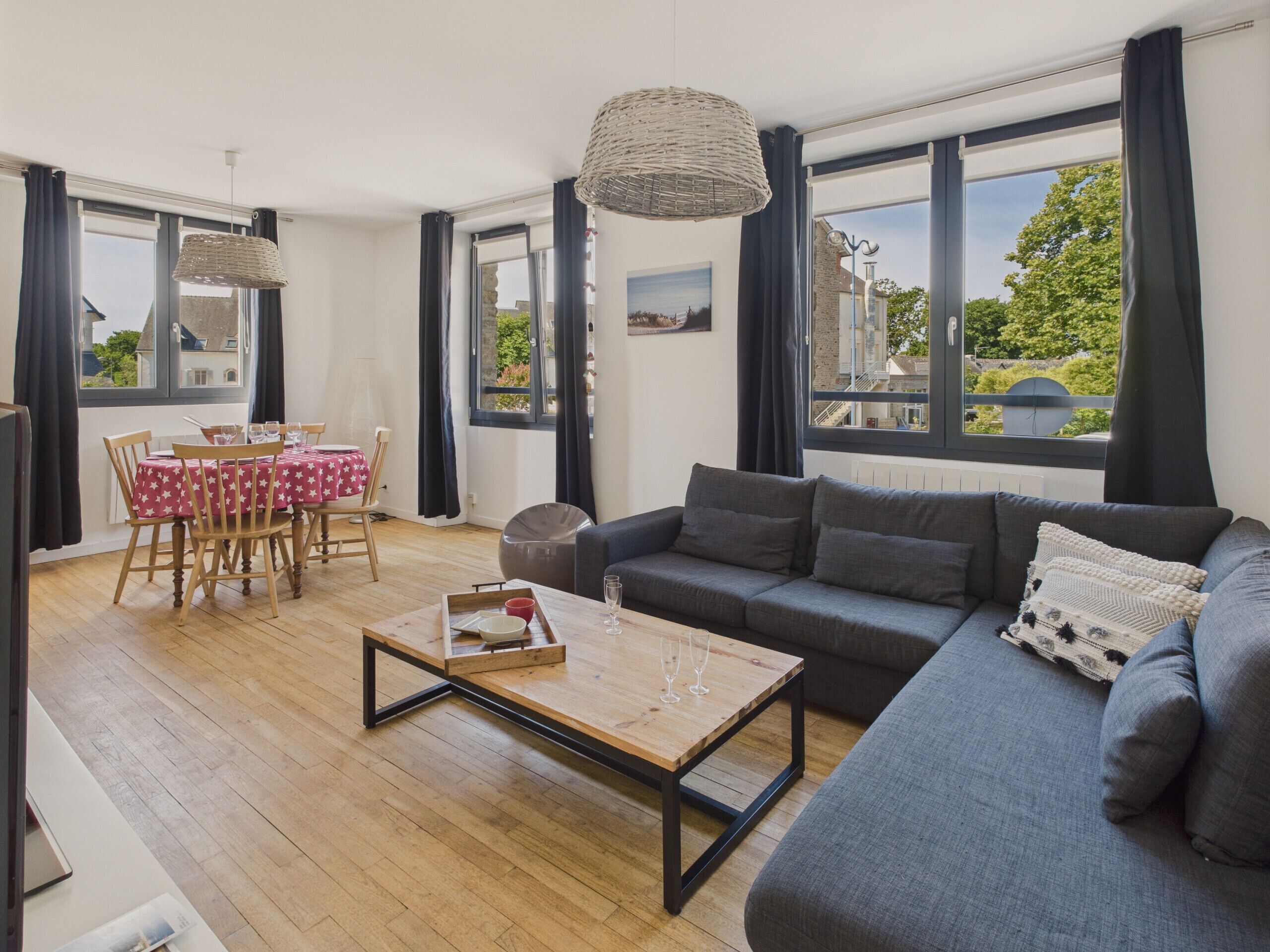 A modern living room with a grey sofa, wooden coffee table, and a dining area with a red star-patterned tablecloth.