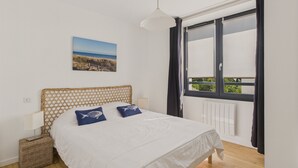 A bedroom with a wooden headboard, white bedding, two pillows with fish designs, a wicker basket, a view of greenery through a window, and a ceiling light.