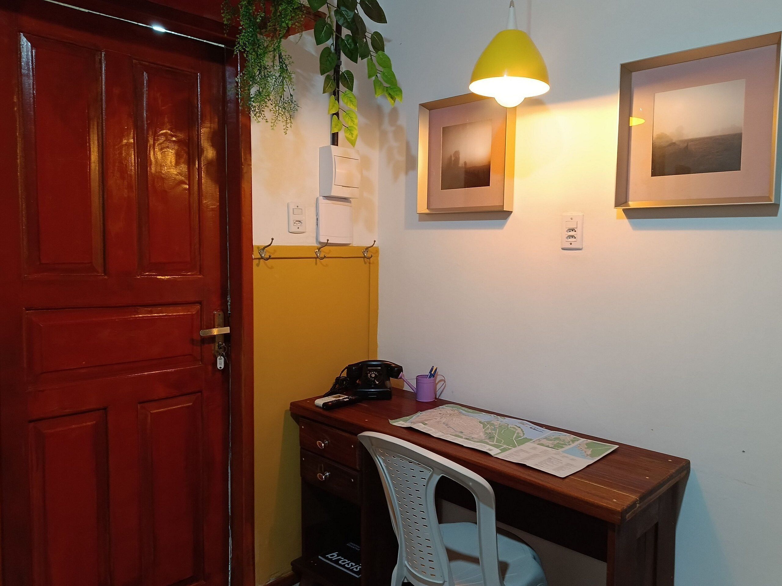 A room with a wooden desk, a chair, a hanging plant, a yellow lamp, and two framed pictures on the wall.