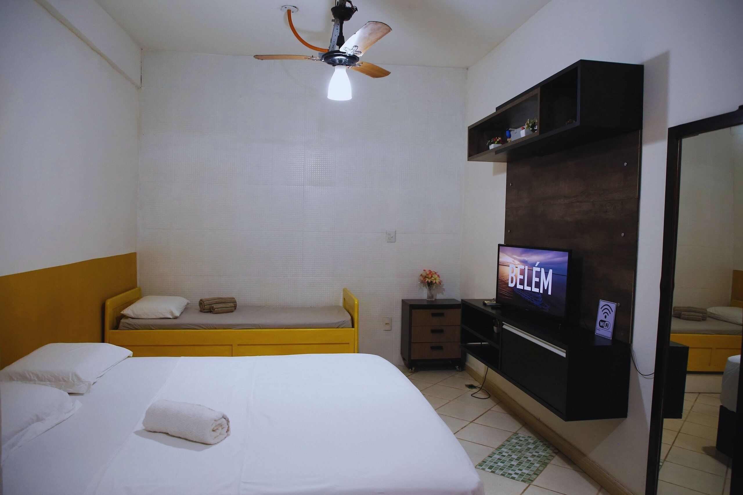 A hotel room with a bed, a TV, a ceiling fan, and a small desk with a vase of flowers.