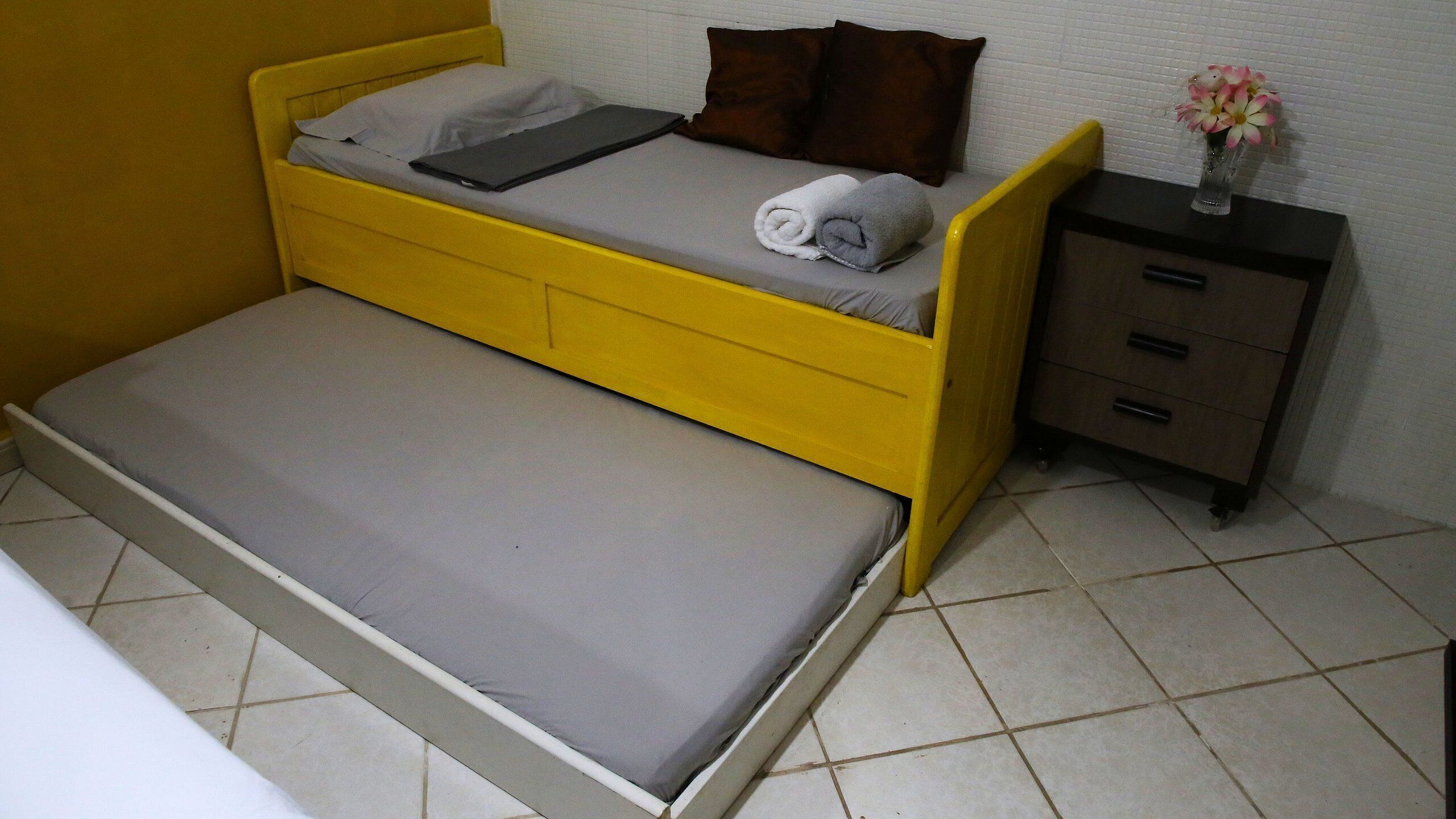 A small, yellow single bed with a grey mattress, a bedside table with drawers, and a vase with flowers on the nightstand.