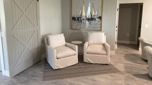 Une pièce comprenant deux fauteuils beiges, une petite table ronde, un tapis à motifs, une grande peinture abstraite et une porte de style rustique.