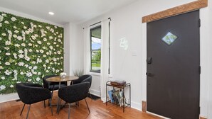 Une pièce avec un plancher en bois, une table ronde entourée de chaises, un arrangement floral fixé au mur, une fenêtre avec des rideaux, une porte sombre dotée d’une petite fenêtre, et une pancarte indiquant « Prenez-y les choses tranquillement ».