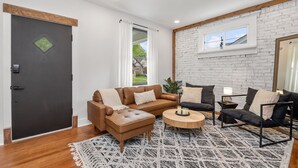 Un salon moderne avec un canapé en cuir marron, un canapé noir et une table basse ronde. On y trouve une fenêtre avec des rideaux blancs, un mur en briques blanches et une poutre en bois au plafond.