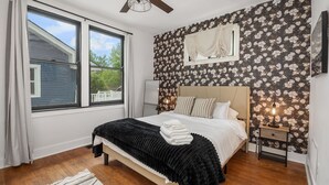 Une chambre à coucher comprenant un lit, une table de chevet, un ventilateur de plafond, une fenêtre avec des rideaux et un mur recouvert d’un papier peint à motifs floraux.