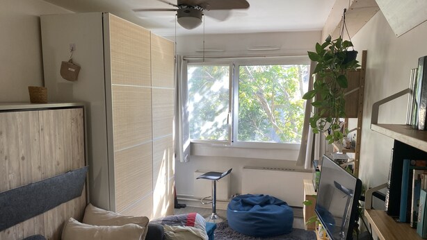 A room with a ceiling fan, a window with a view of trees, a blue bean bag, a desk with papers, and a TV on a wooden stand. - Studio Confortable Paris 13 (Paris)