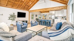 A spacious living area with a high wooden ceiling, a flat-screen TV, and a kitchen with white cabinets and a bar area. - Park Place: Luxury Riverfront Home, Walk to Lake Michigan & Chain Ferry, Deck (Saugatuck)