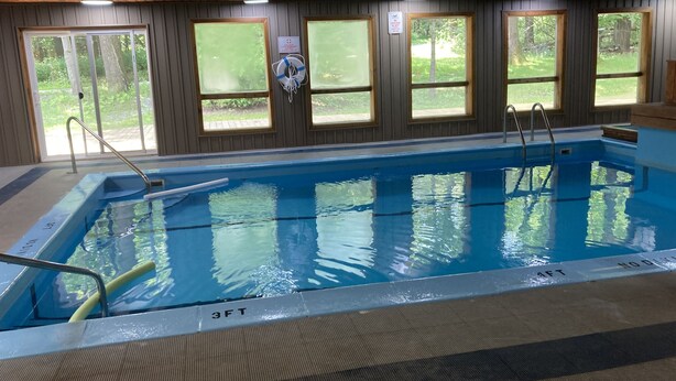 An indoor swimming pool with clear water, wooden walls, and windows offering views of greenery.