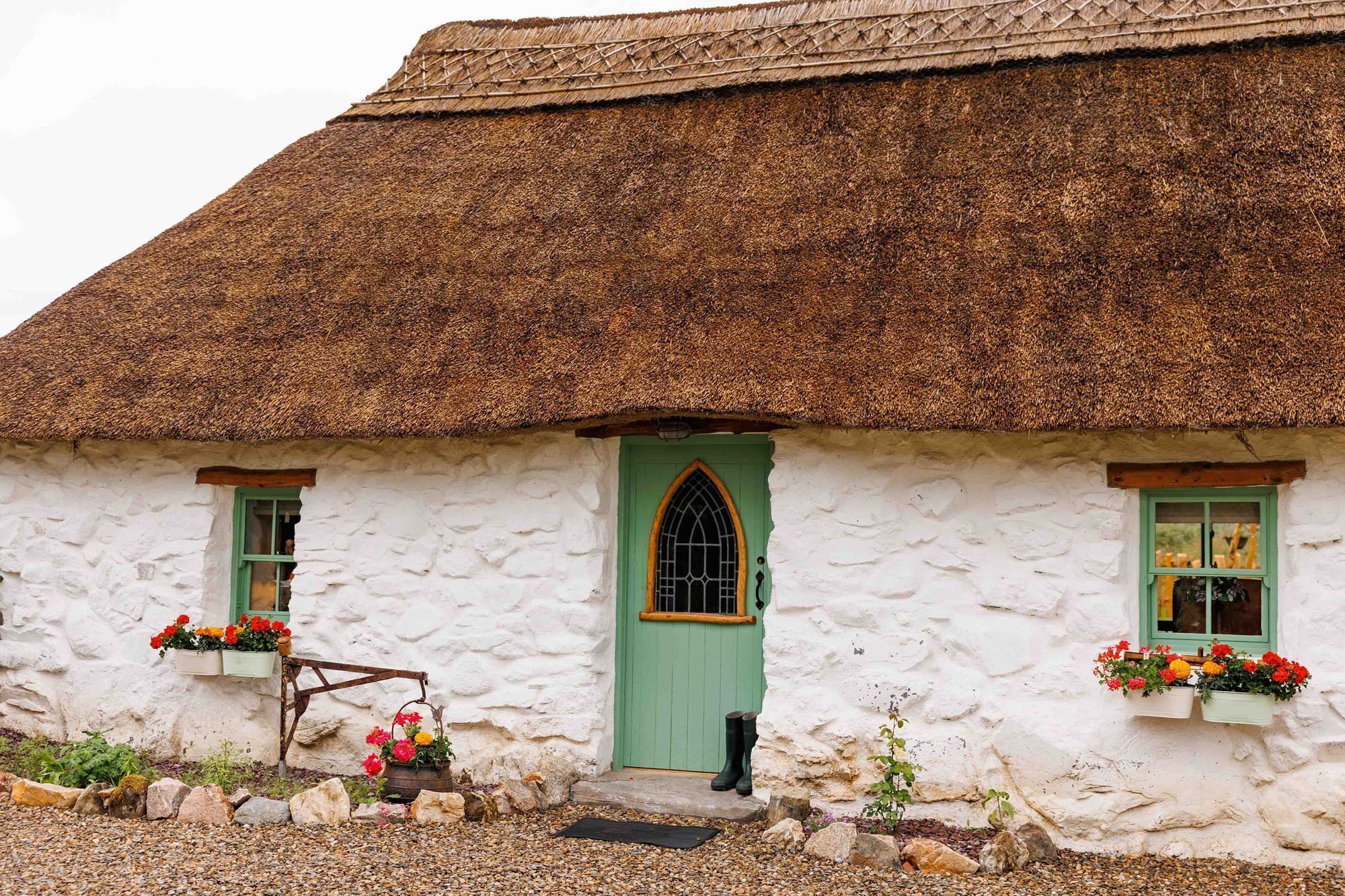 A traditional thatched-roof cottage with a green door, white walls, and flower boxes on the windowsills.
