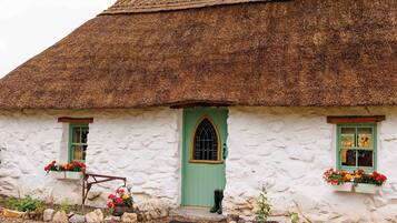 A traditional thatched-roof cottage with a green door, white walls, and flower boxes on the windowsills.