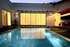 Una piscina con borde de baldosas, puerta corrediza de vidrio y un amplio ventanal con cortinas.