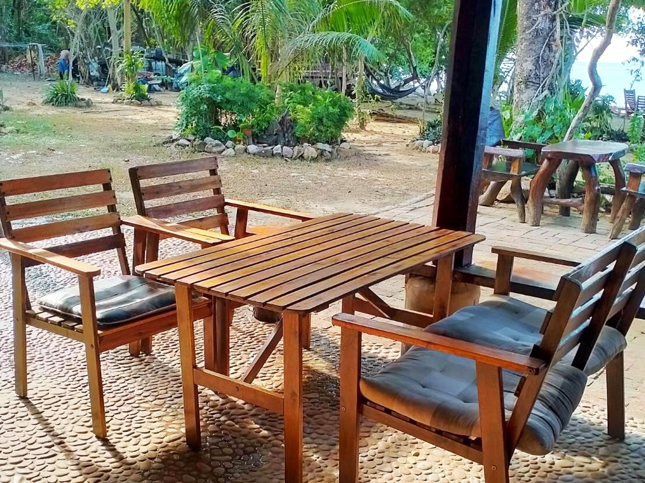 A wooden table and chairs set outdoors with a view of a tropical garden and beach.
