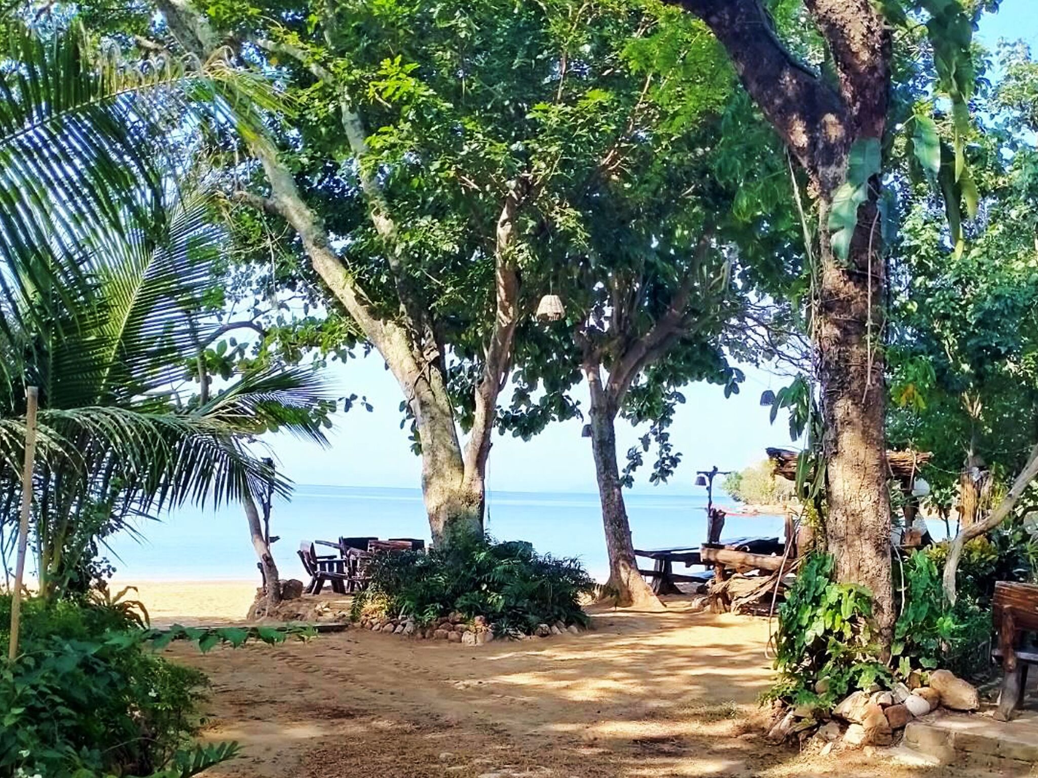 A beach path leading to a wooden deck with chairs and a table, surrounded by trees and vegetation.