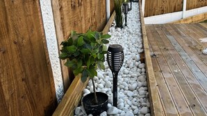 Una terraza de madera con guijarros blancos, una maceta negra con una planta verde y un foco de jardín negro.