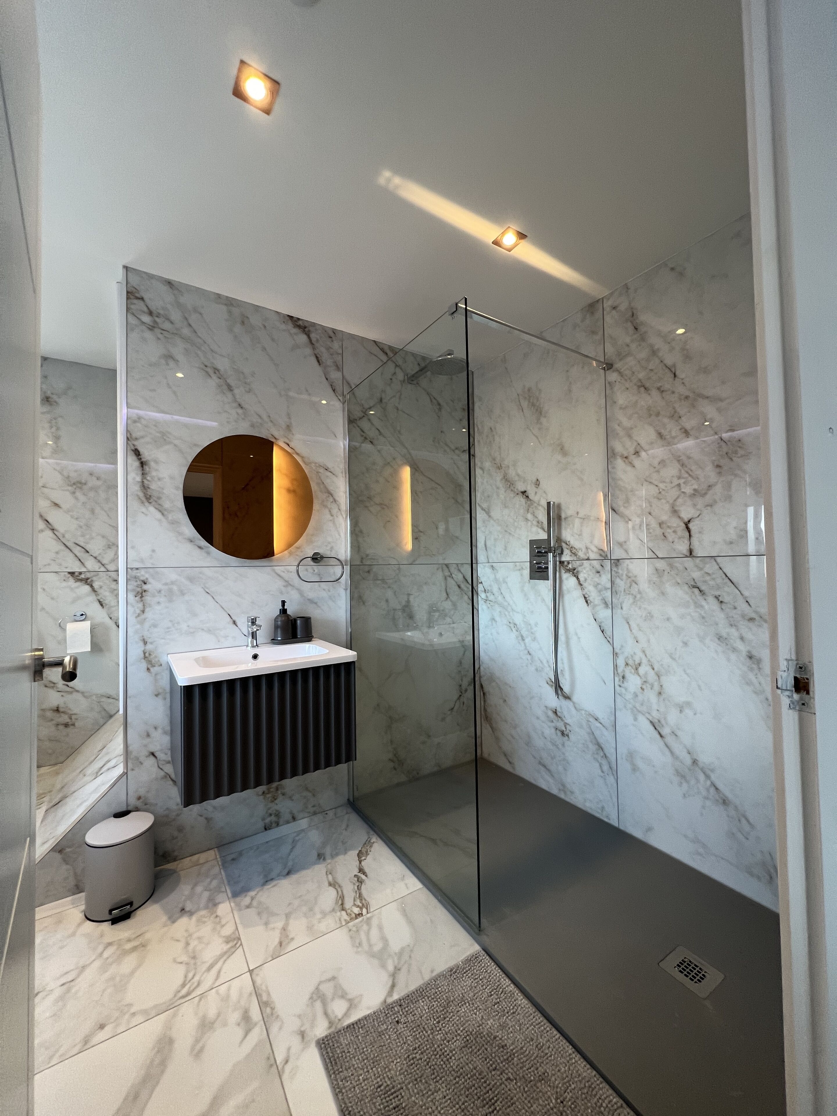 Un bagno moderno con box doccia in vetro, pavimento in marmo e specchio tondo sopra il lavabo.